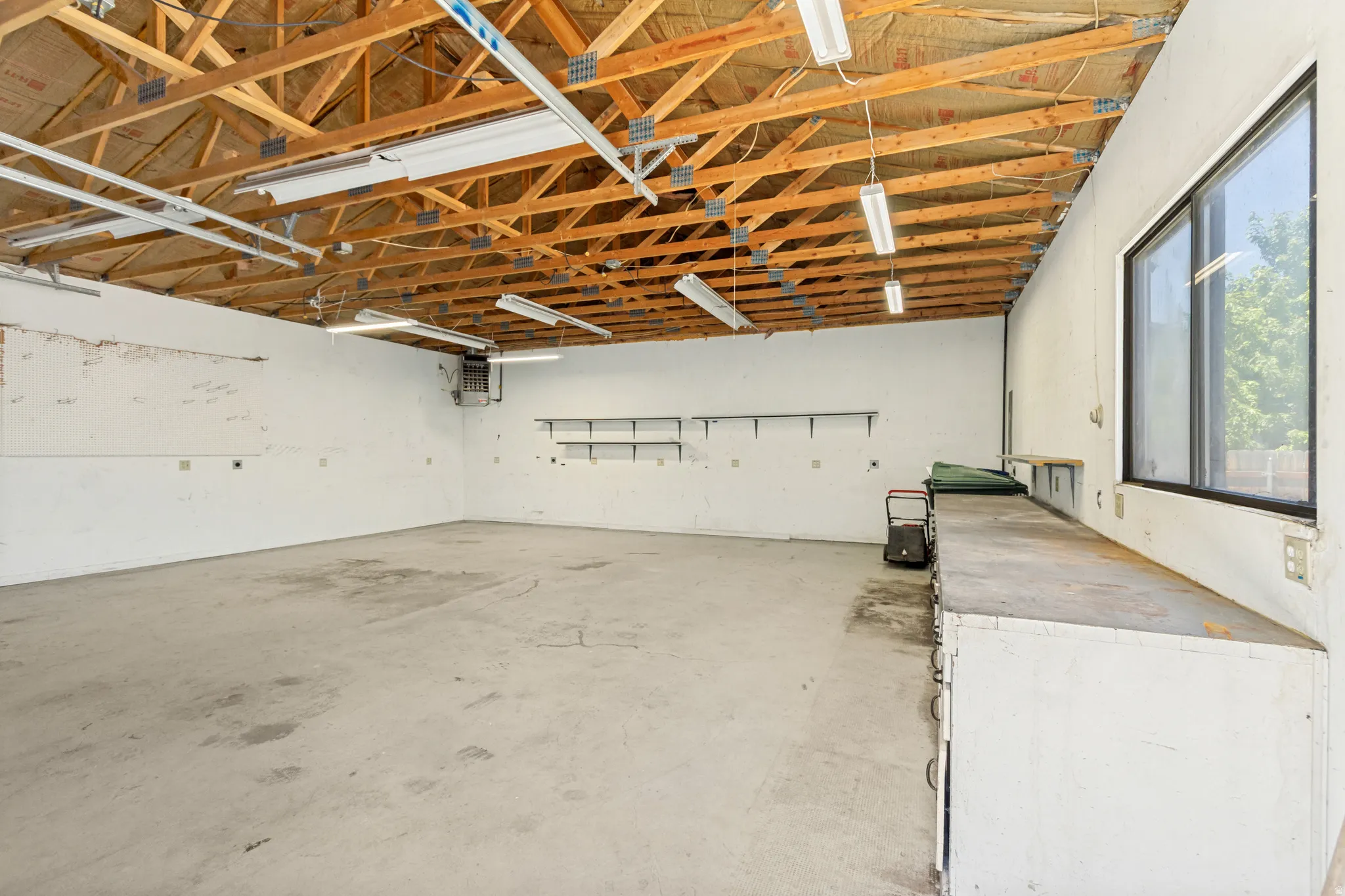 Spacious interior featuring exposed wooden rafters, concrete flooring, fluorescent lighting, a large window, and built-in shelving and workbench.