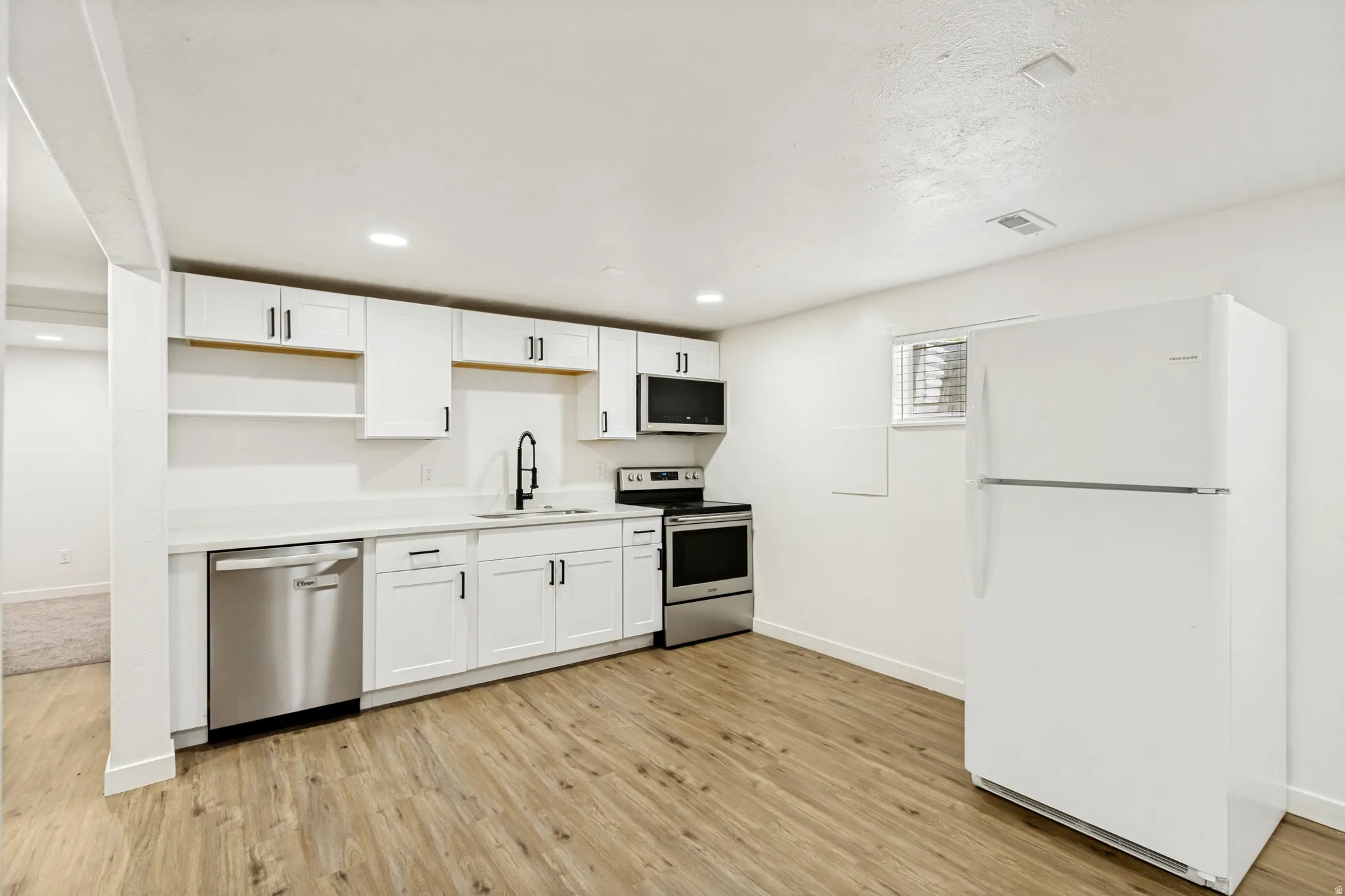 Full basement kitchen featuring white cabinetry with black hardware, stainless steel appliances, light-toned wood-finish flooring, and a white refrigerator