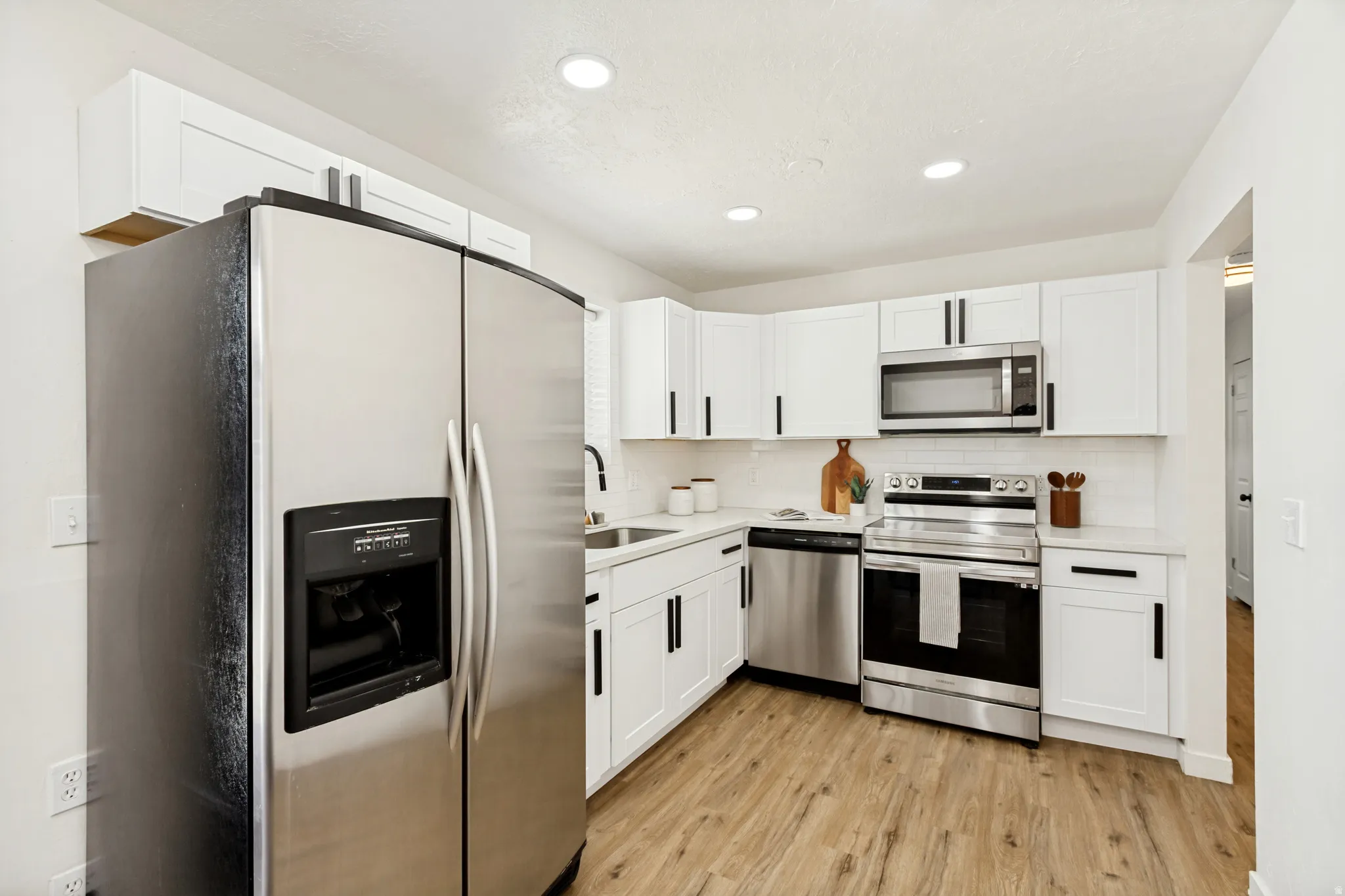 Modern kitchen featuring stainless steel appliances, white cabinetry with matte black hardware, light-toned wood-look flooring, and recessed lighting