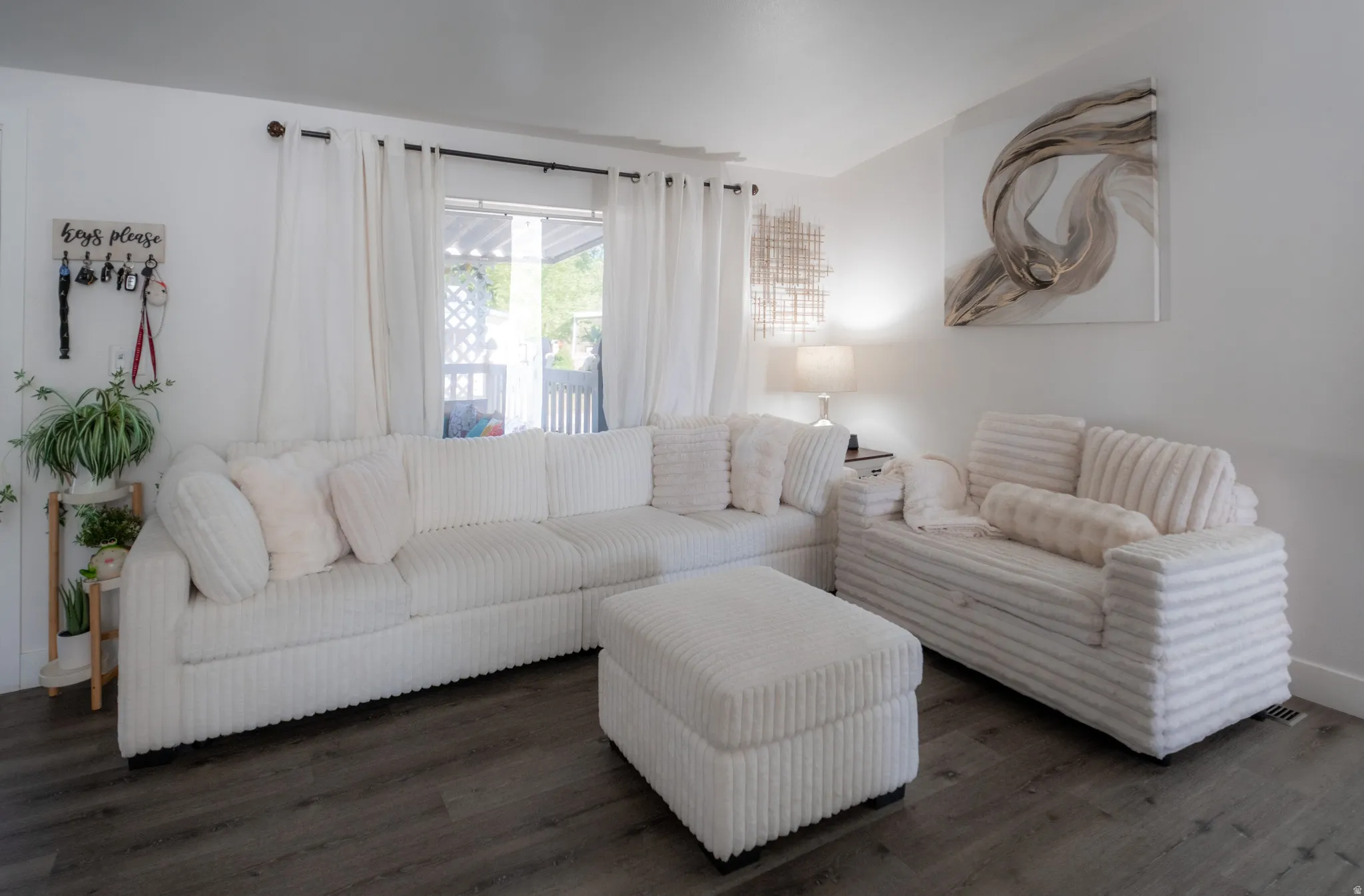 Bright living area featuring wood-finish flooring, white walls, and a large window with white curtain panels