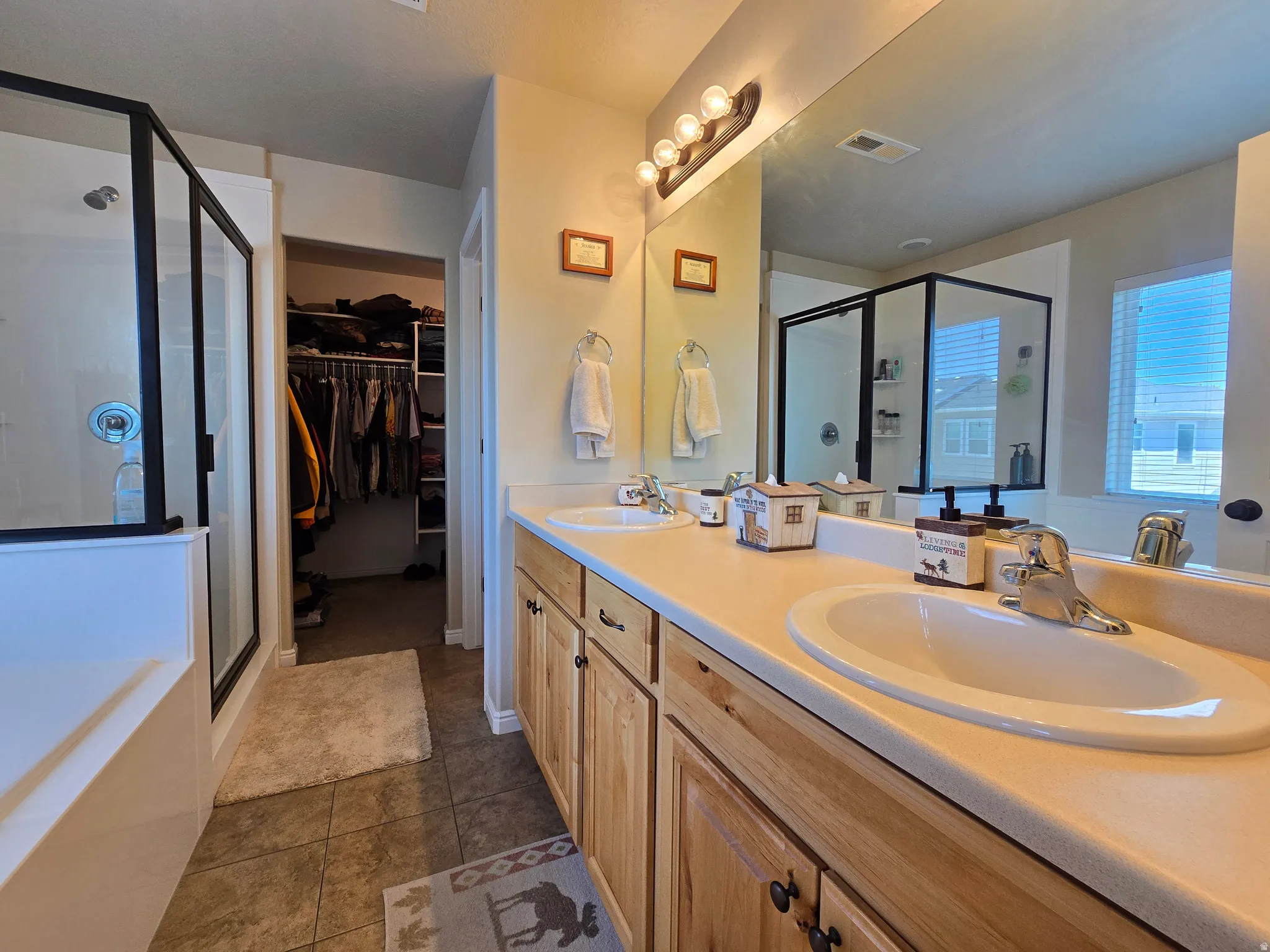 Bathroom featuring a double vanity with light wood cabinetry and integrated sinks, a full-width mirror, and a walk-in shower with a black-framed glass enclosure