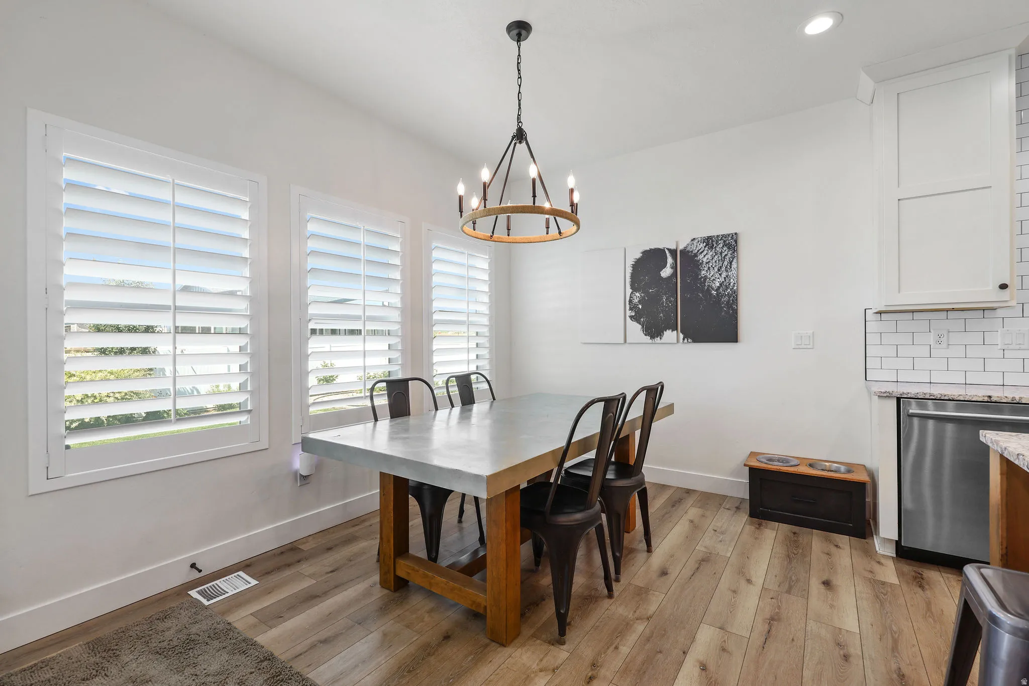 Dining area featuring light wood-finish flooring, white plantation shutters, a dark metal chandelier, and a kitchen area with white subway tile backsplash and stainless steel appliances