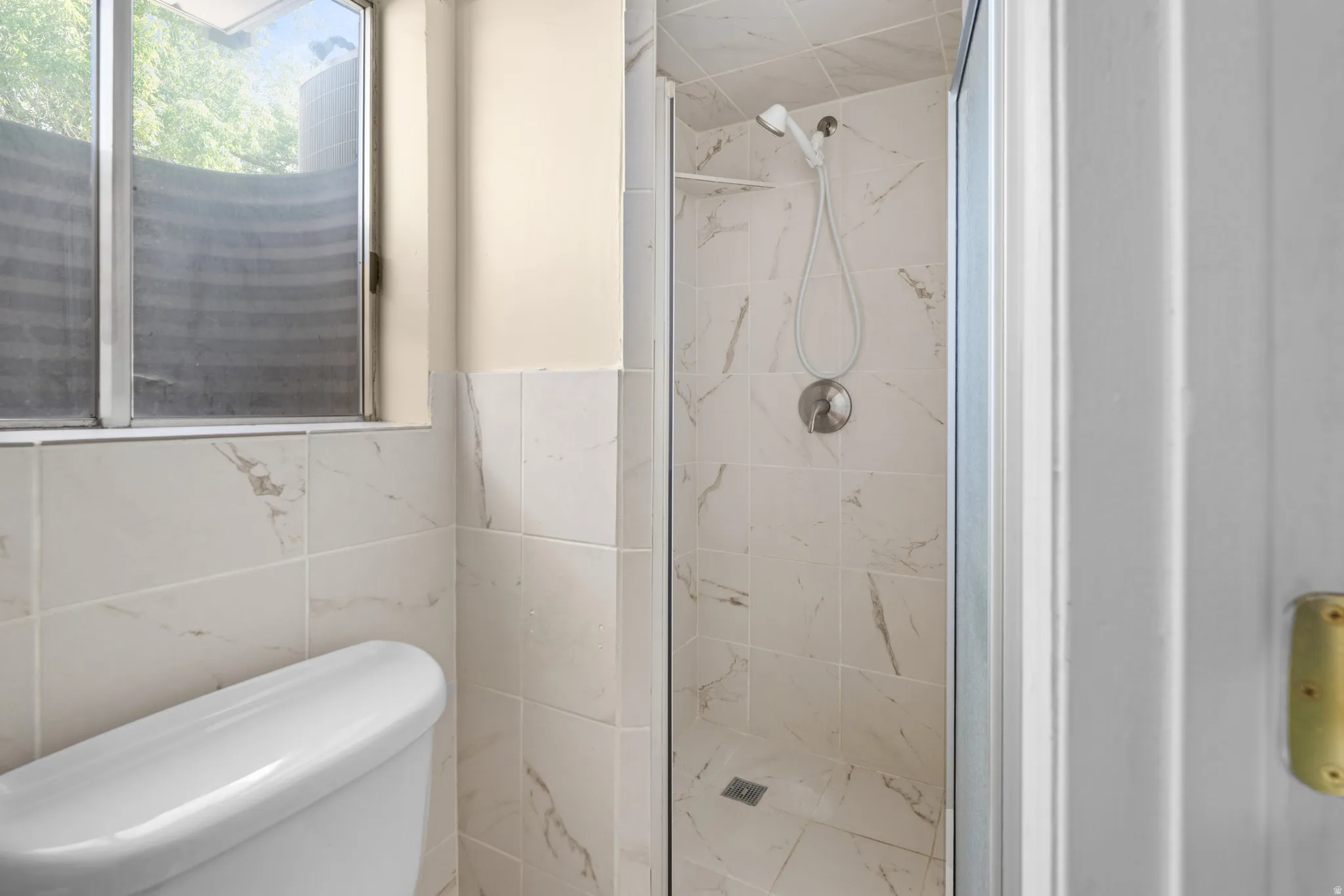 Shower enclosure with floor-to-ceiling marbled-patterned tiling, a handheld shower fixture, and a built-in corner shelf