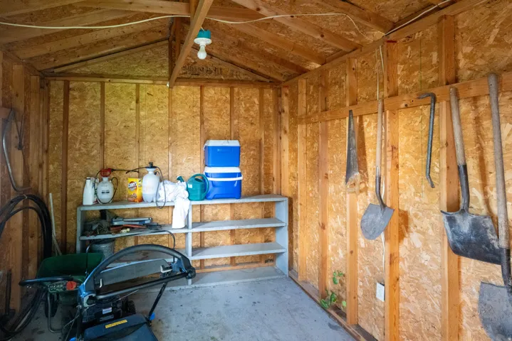 Utility shed featuring wood paneling, open rafter ceiling, built-in shelving, and interior lighting