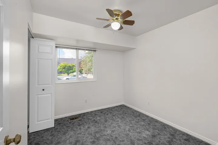 Interior room featuring a ceiling fan with wood-finish blades, a dual-pane window, white baseboards, and dark gray patterned carpeting