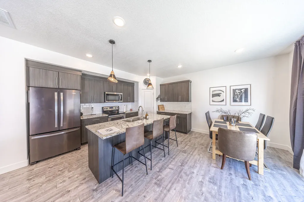 Open-concept kitchen and dining area featuring wood-finish flooring, dark wood-tone cabinetry, stainless steel appliances, a subway tile backsplash, and a large central island with pendant lighting