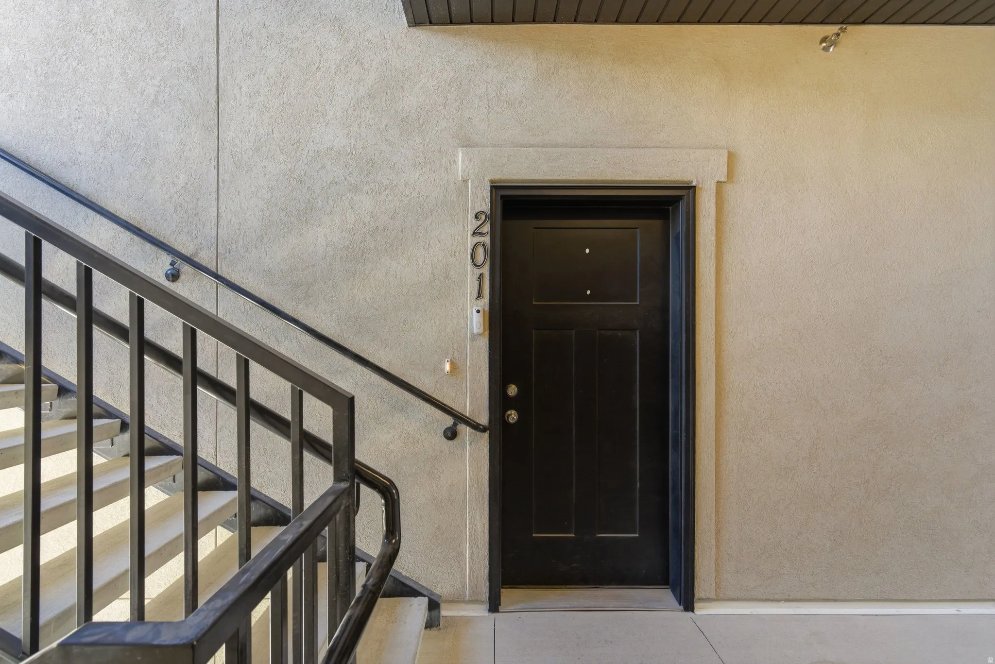 Exterior walkway featuring a substantial black paneled entry door with surrounding trim, unit numbering, and a durable metal railing bordering a concrete staircase