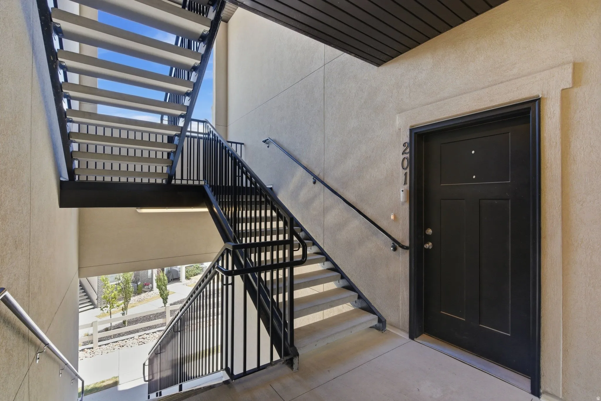 Exterior common area featuring multi-level open-riser stairs with black metal railings, textured stucco walls, and a black paneled entry door