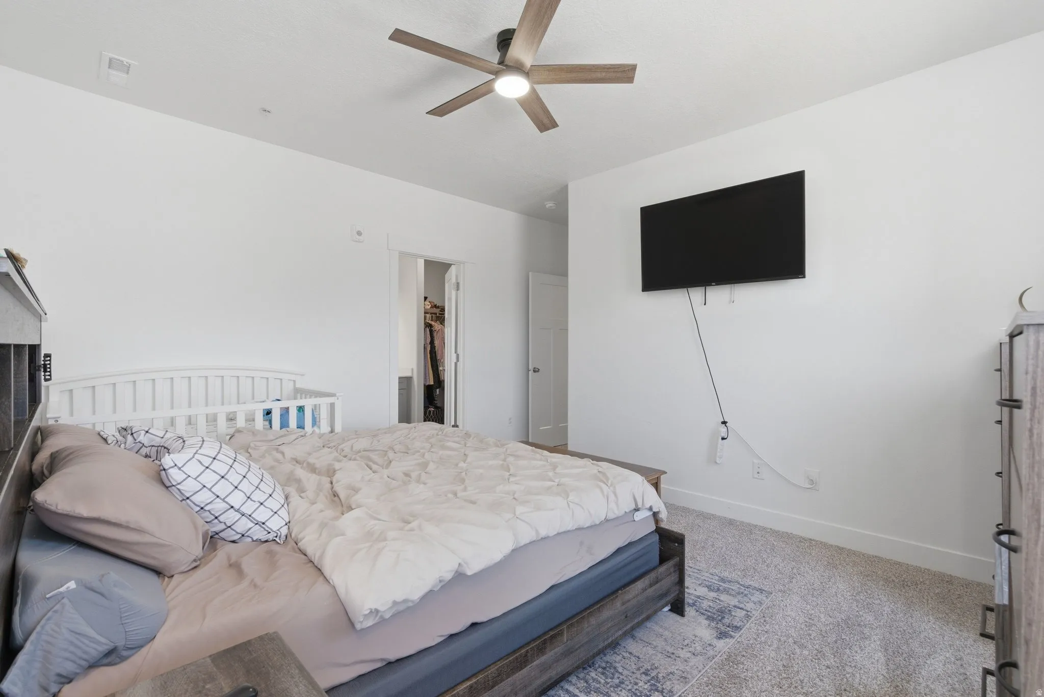 Spacious bedroom featuring light-toned carpeting, a ceiling fan with integrated lighting, and a wall-mounted television bracket