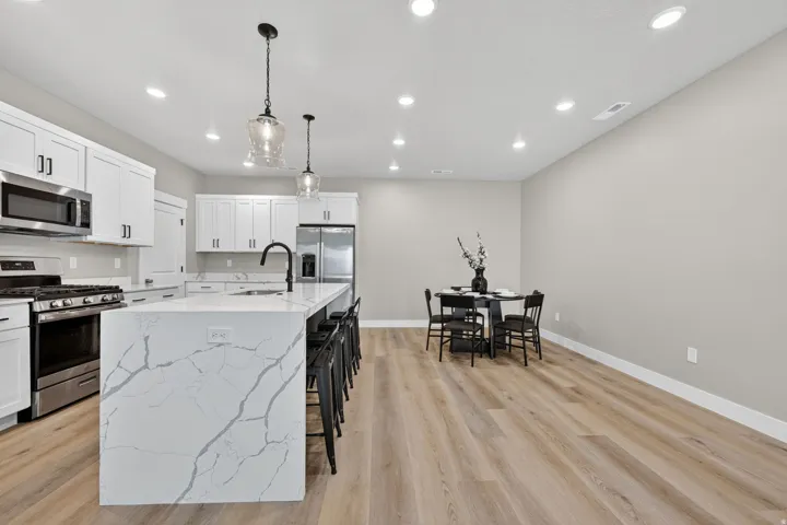 Spacious kitchen featuring an oversized island with waterfall-edge quartz countertops, stainless steel appliances, white shaker cabinetry, and wood-finish flooring
