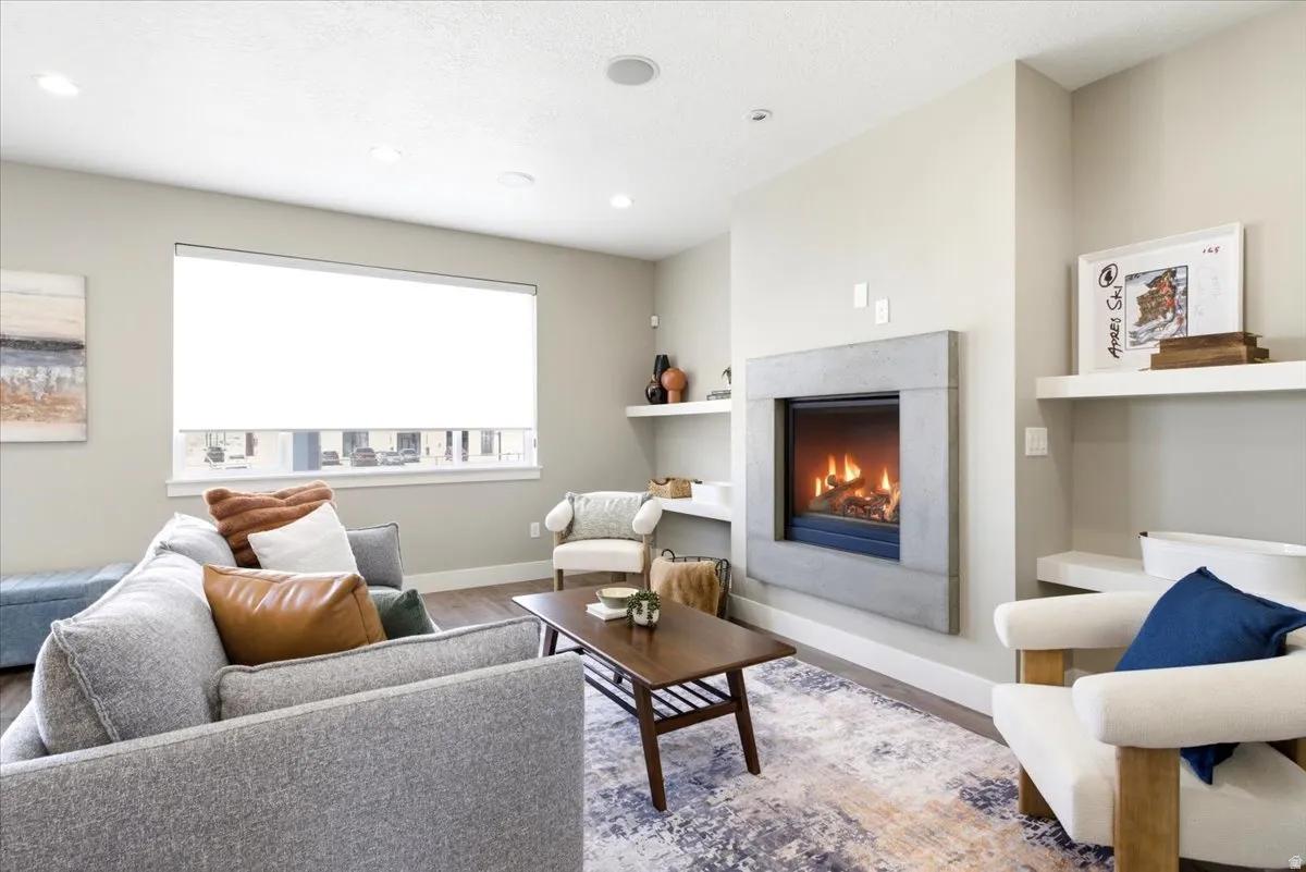 Living area featuring a modern wall-mounted fireplace with a concrete surround, wood-finish flooring, recessed lighting, a large window with roller shades, and built-in floating shelves