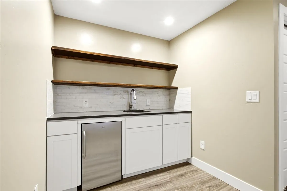 Compact wet bar featuring white cabinetry, a stainless steel under-counter refrigerator, a stainless steel sink with a gooseneck faucet, and a stone-look tile backsplash
