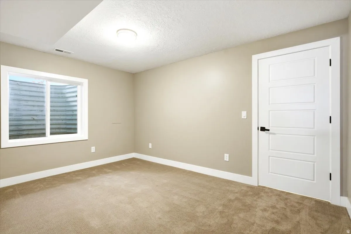 Carpeted room featuring a white-framed egress window, light beige wall paint, white baseboards, a flush-mount ceiling light fixture, and a white paneled door with a black handle