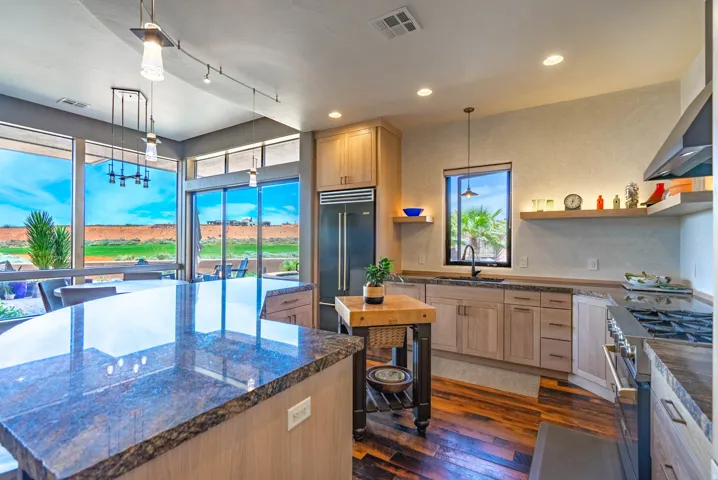 Spacious kitchen featuring an expansive granite island, wood-finish flooring, light wood cabinetry, stainless steel appliances, and large windows with outdoor access