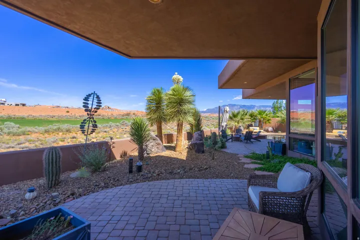 Expansive covered patio featuring paver flooring, a desert landscape with mature palms and cacti, and a full-height glass exterior wall