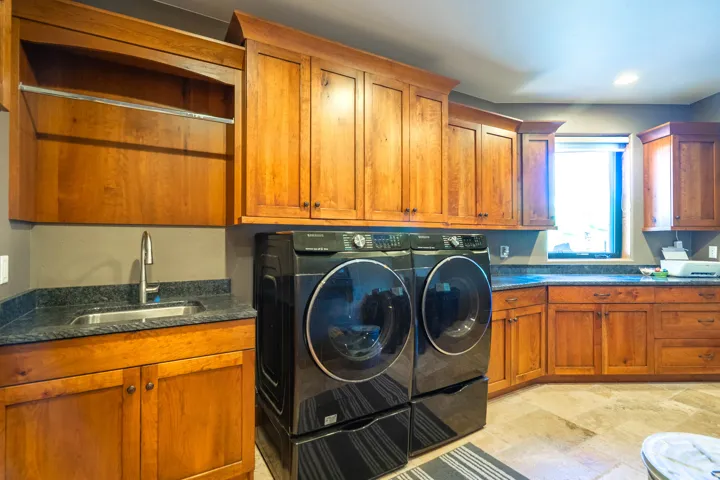 Dedicated laundry space featuring a full-size washer and dryer set, extensive wood cabinetry, dark stone countertops, a stainless steel utility sink, and tile flooring