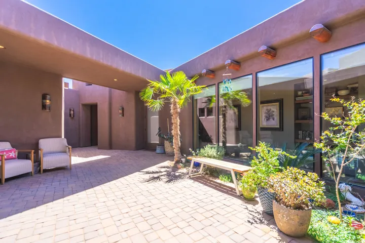 Paved outdoor courtyard featuring a central palm tree, surrounding garden beds, multiple large window panels, and a covered walkway with recessed lighting