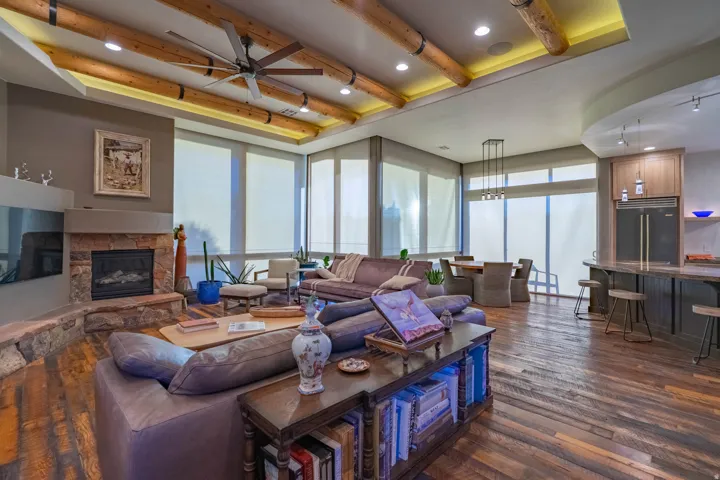 Open-concept living space featuring wood-finish flooring, a stone-faced fireplace with hearth, exposed wood ceiling beams with recessed lighting, and a kitchen island with bar seating