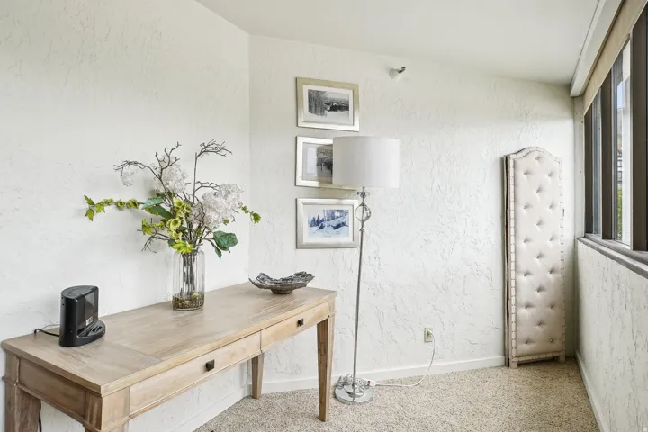 Light-toned wood-finish console table with two drawers, textured white walls, light-colored carpet flooring, and a bank of windows providing natural light