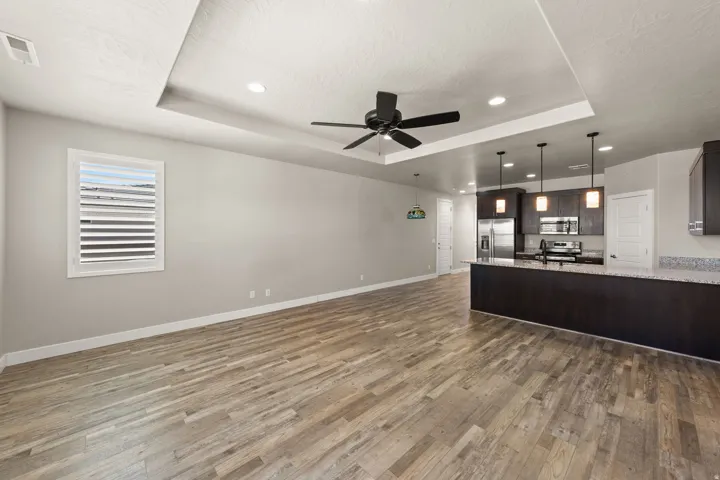 Open-concept living area featuring wood-finish flooring, a tray ceiling with a ceiling fan, recessed lighting, and a modern kitchen with dark cabinetry, stainless steel appliances, and a peninsula with a stone-finish countertop