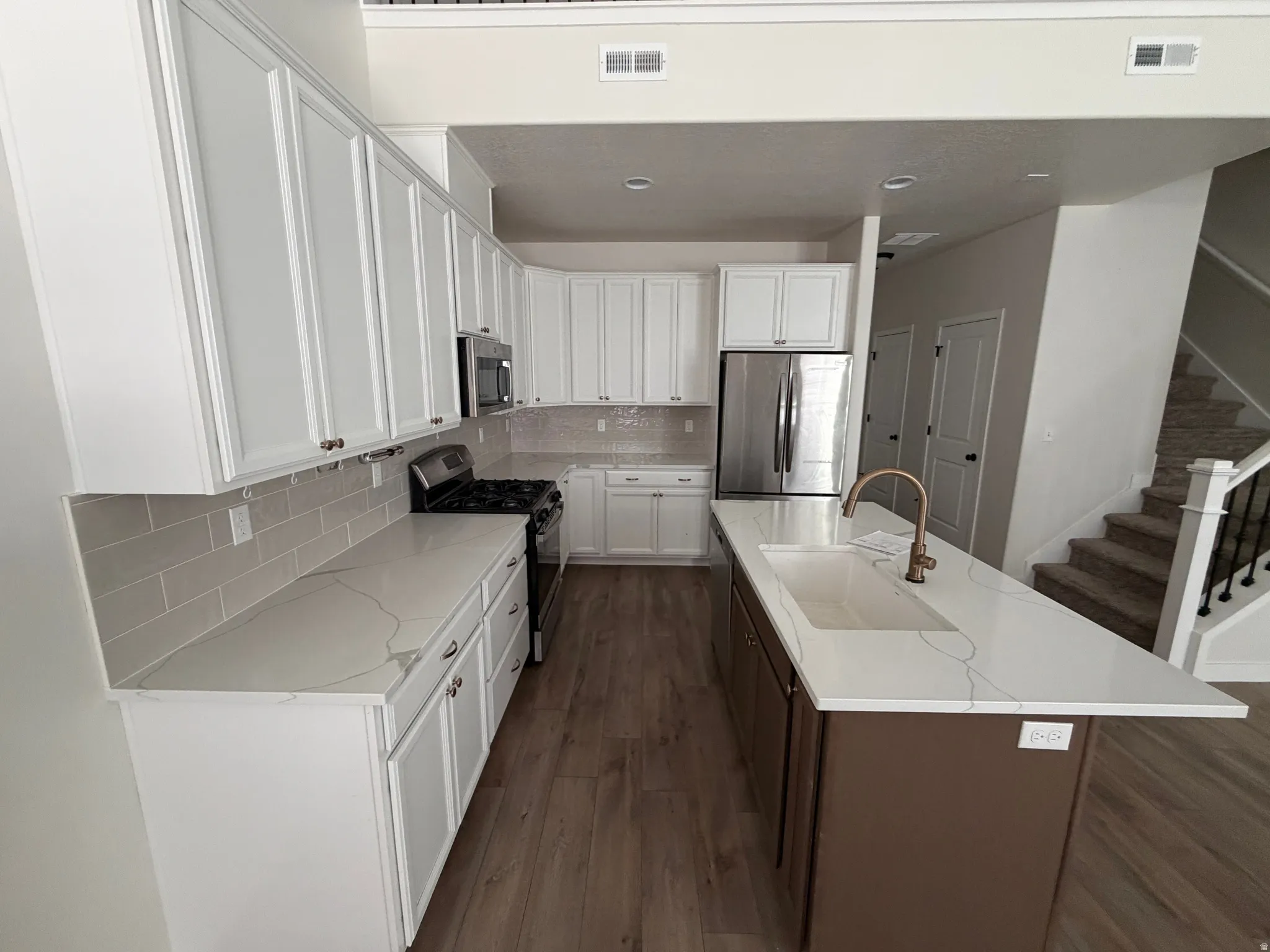 Contemporary kitchen featuring white shaker cabinetry, light-toned countertops with subtle veining, stainless steel appliances, a subway tile backsplash, and a central island with an integrated sink and bronze-finish faucet