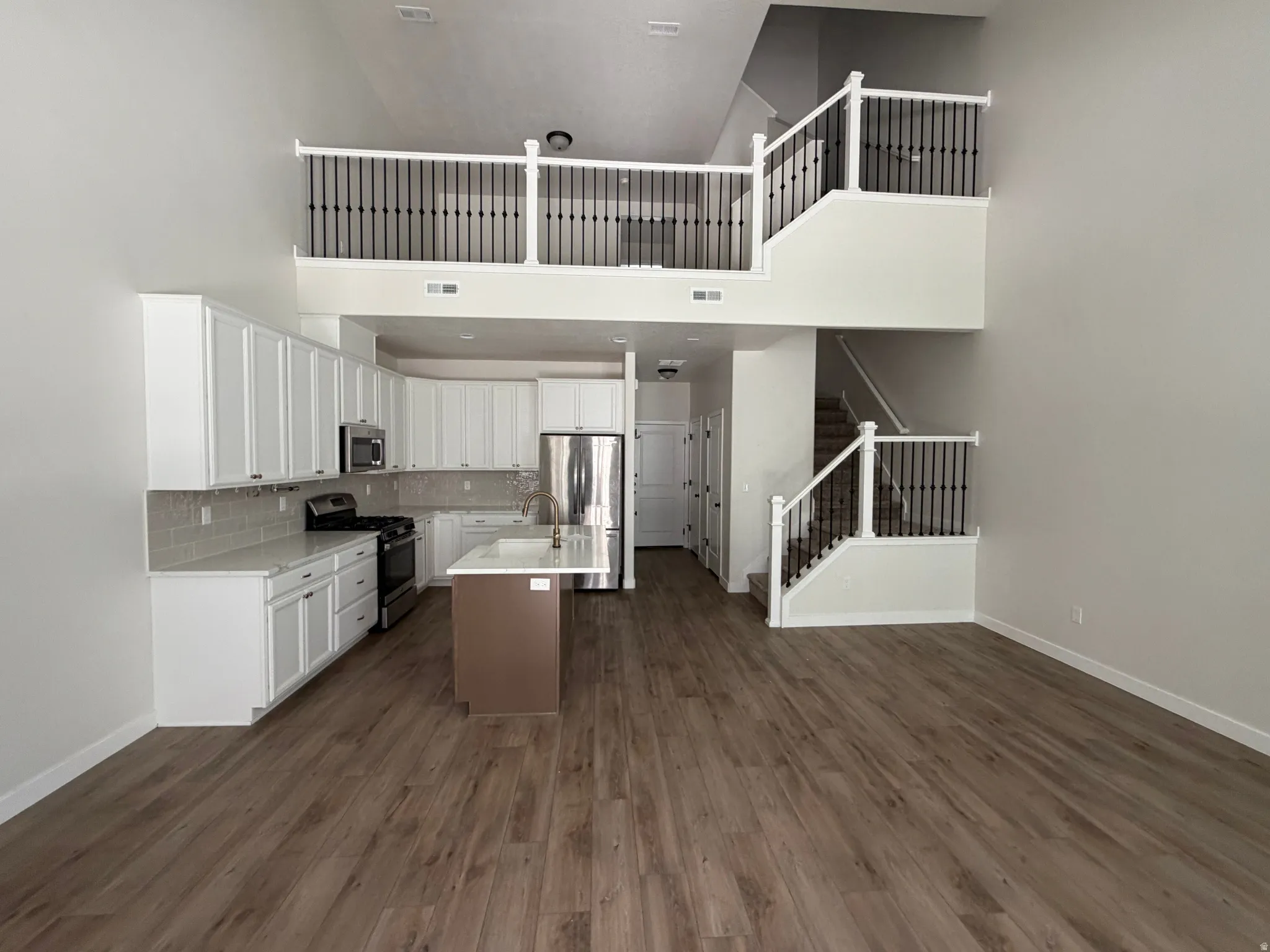 Open-concept living area featuring high ceilings, wood-finish flooring, a contemporary kitchen with white cabinetry, stainless steel appliances, and a central island, and a staircase leading to an upper-level loft