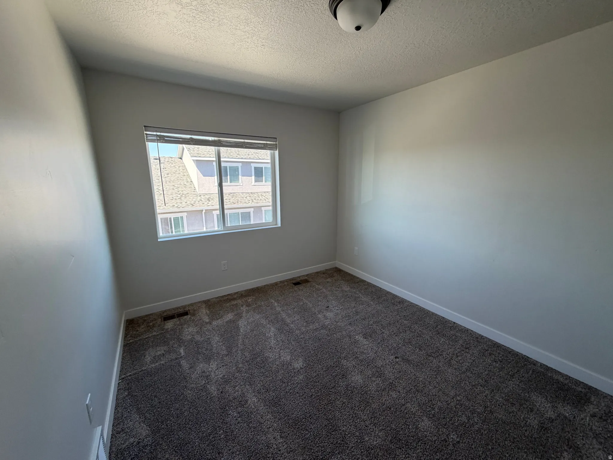 Room featuring a window with blinds, a ceiling-mounted light fixture, light gray walls, white baseboards, and dark gray patterned carpet
