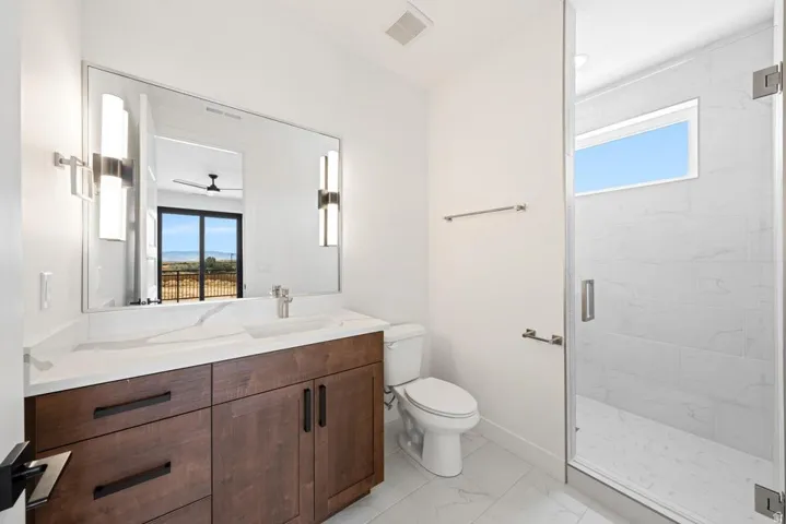 Modern bathroom featuring a wood-finish vanity with a white countertop and rectangular sink, a large wall-mounted mirror with integrated lighting, a glass-enclosed shower with white tiled walls, and light-colored tile flooring