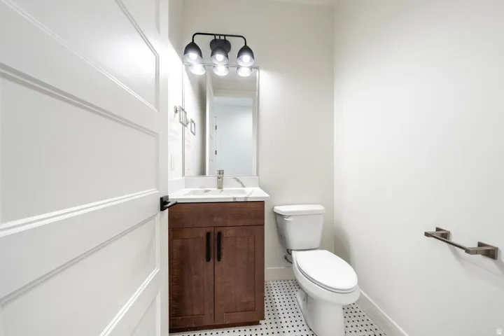 Powder room featuring a white paneled door with black hardware, a vanity with a dark wood-finish cabinet and a light-toned countertop, a rectangular mirror, a three-light vanity fixture with black accents, and small black and white mosaic floor tiles