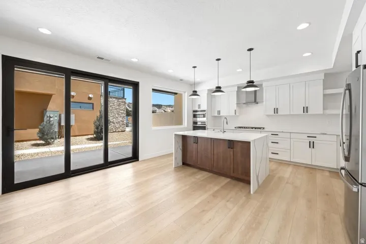 Open-concept kitchen featuring wood-finish flooring, white cabinetry, integrated stainless steel appliances, a center island with a waterfall countertop and contrasting wood-finish base, and recessed lighting with pendant fixtures