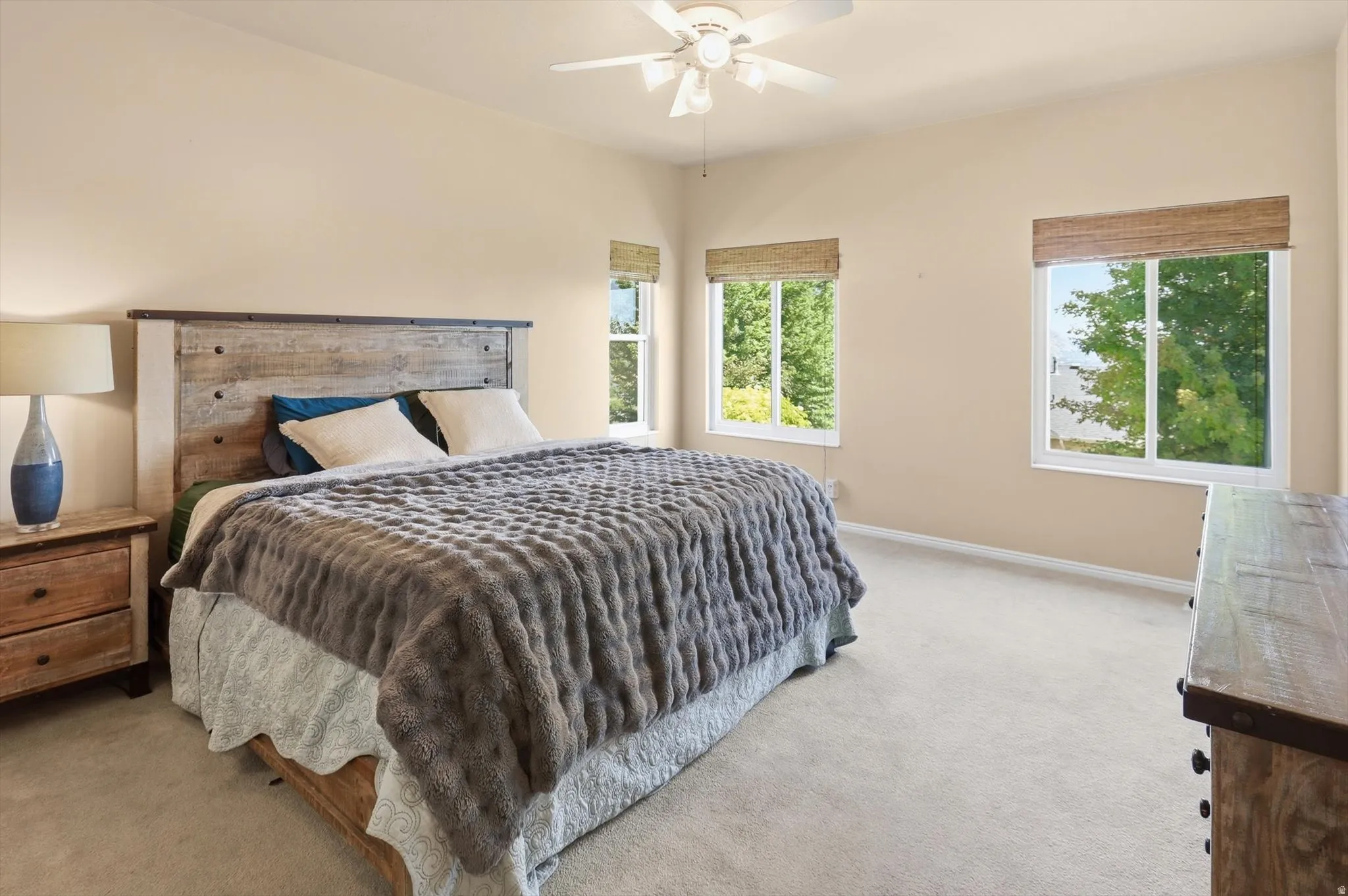 Carpeted bedroom featuring a rustic wood headboard, a ceiling fan, and two windows with natural woven shades