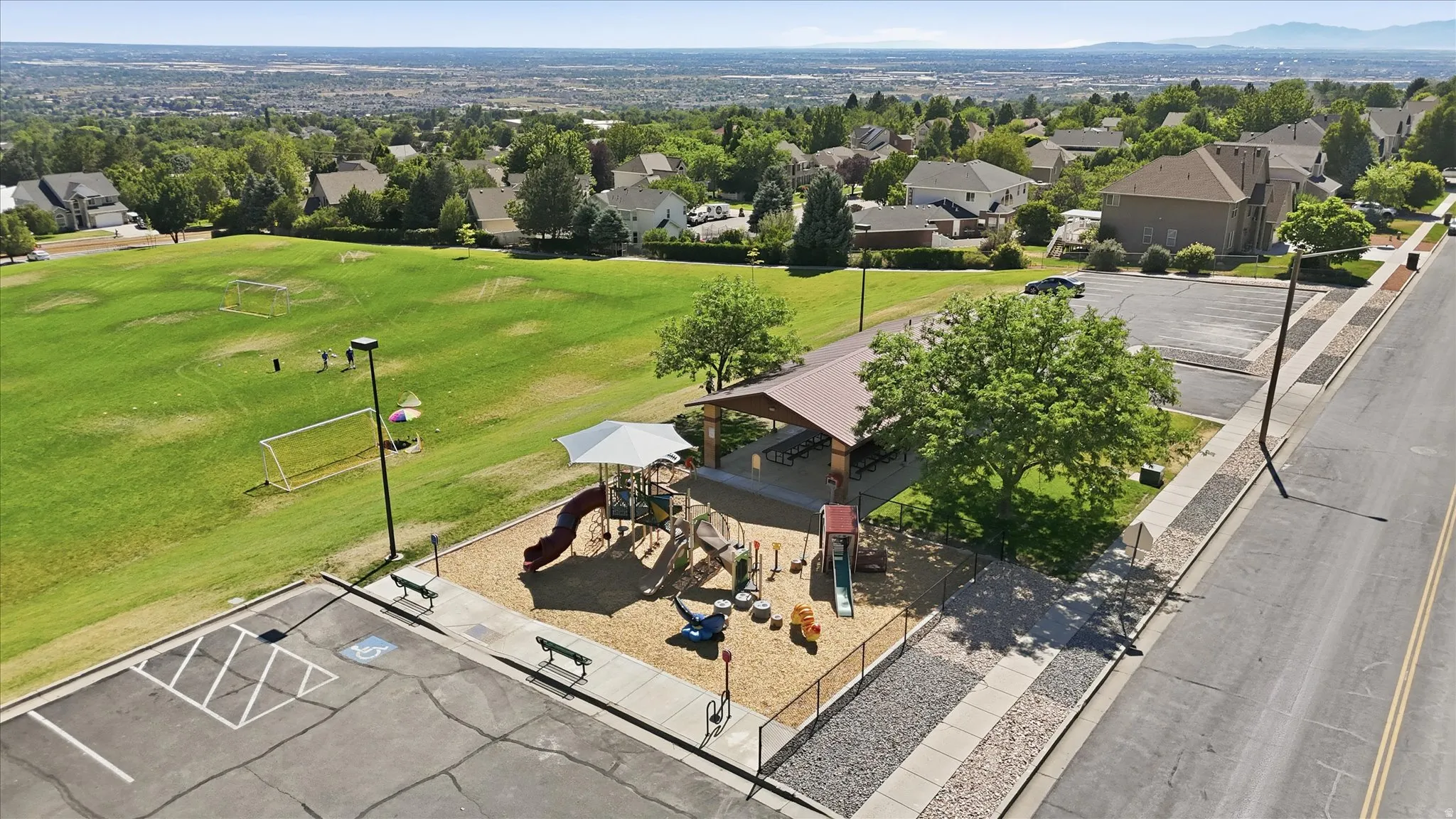 Aerial perspective showcasing a community park with a large green field, a dedicated playground area, a covered picnic pavilion, and a paved parking lot