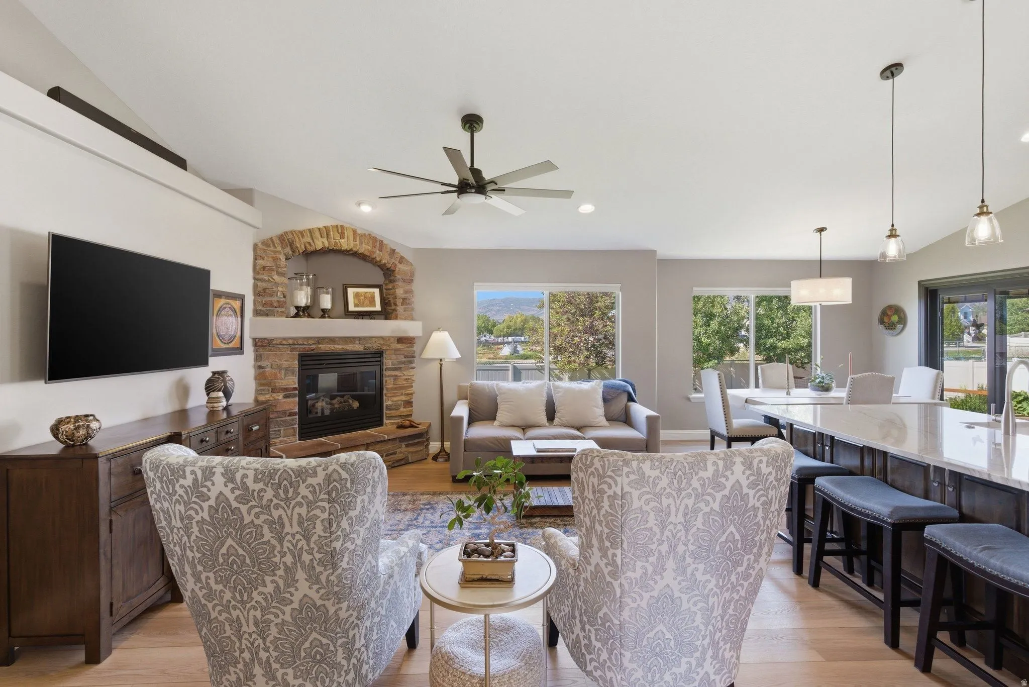 Vaulted ceiling living area featuring a stone-clad fireplace with an arched opening, wood-finish flooring, a ceiling fan, and large windows