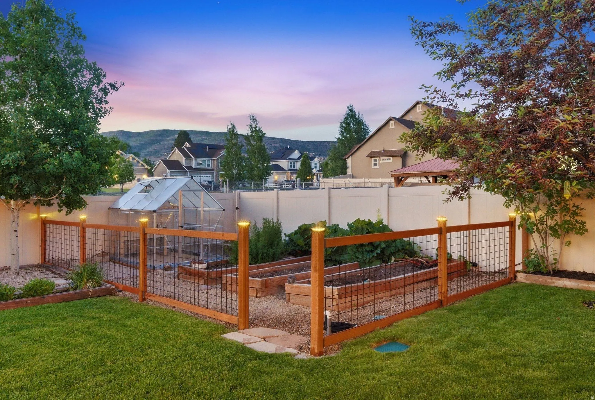 Backyard featuring a functional greenhouse, raised garden beds, wood and wire fence, manicured lawn, and mature trees