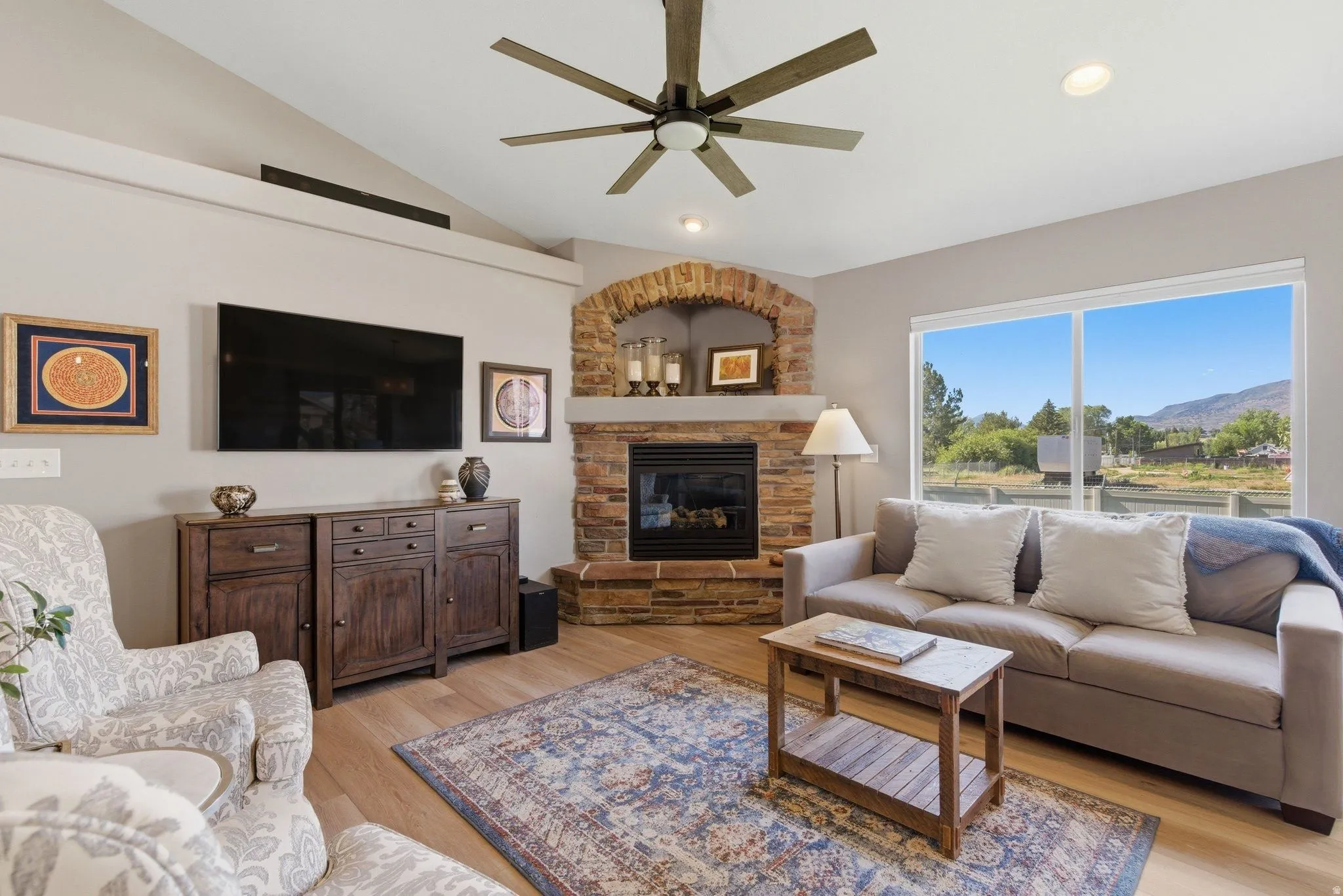 Living area featuring a floor-to-ceiling stone fireplace with an arched brick accent, light wood-finish flooring, and a vaulted ceiling with a ceiling fan