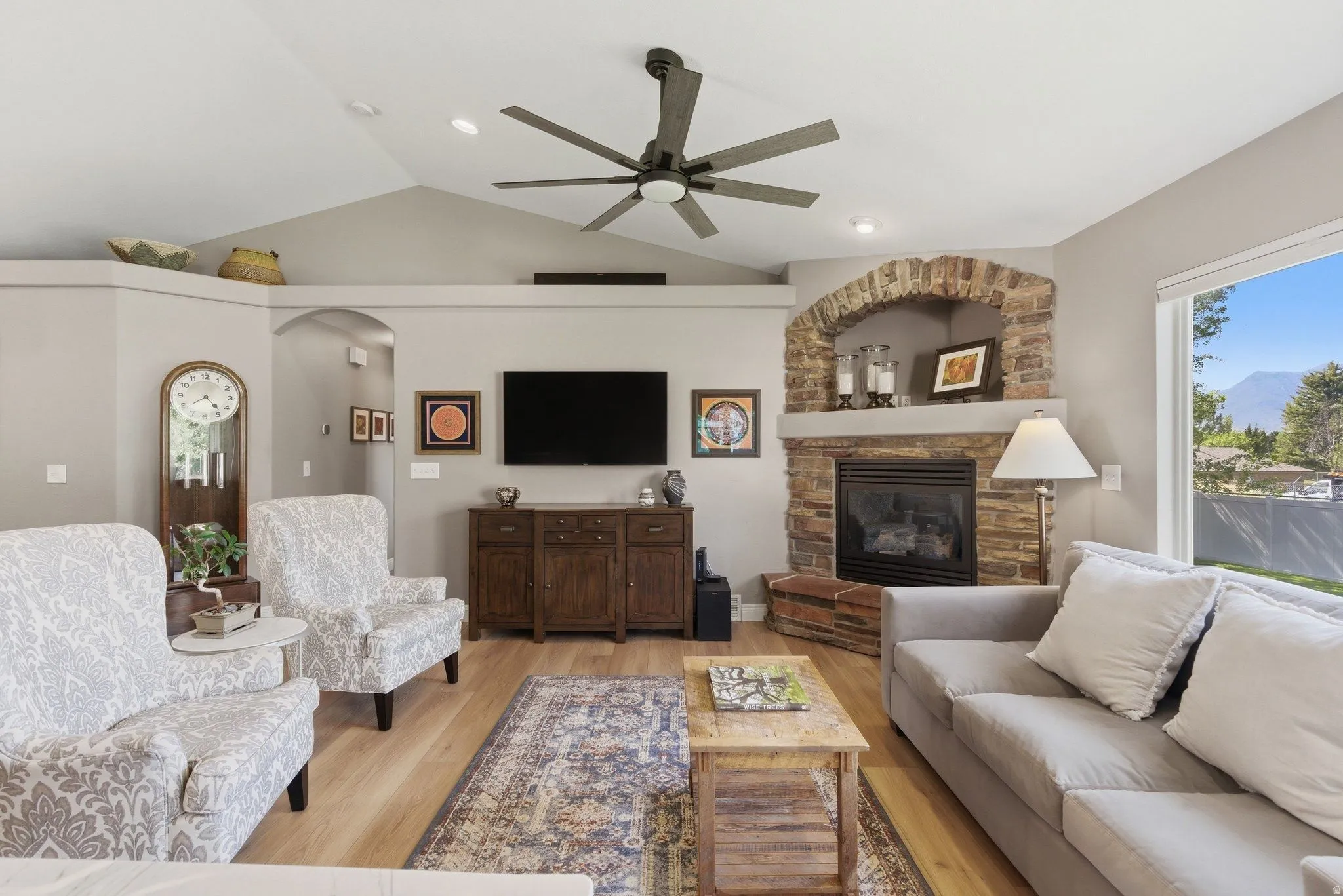Vaulted ceiling living area featuring a stone fireplace with hearth, wood-finish flooring, recessed lighting, and a large picture window
