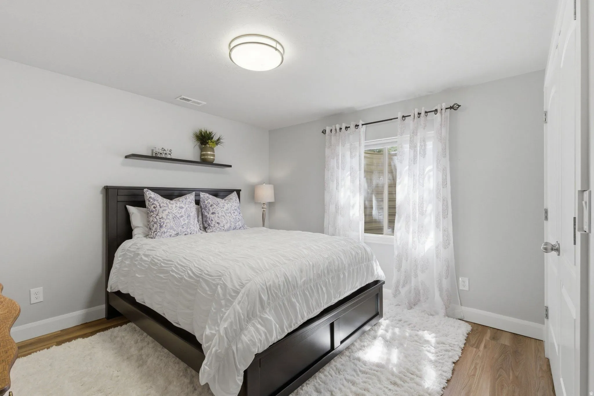 Bedroom featuring wood-finish flooring, light gray walls, a window with sheer white curtains, and a flush-mount ceiling light
