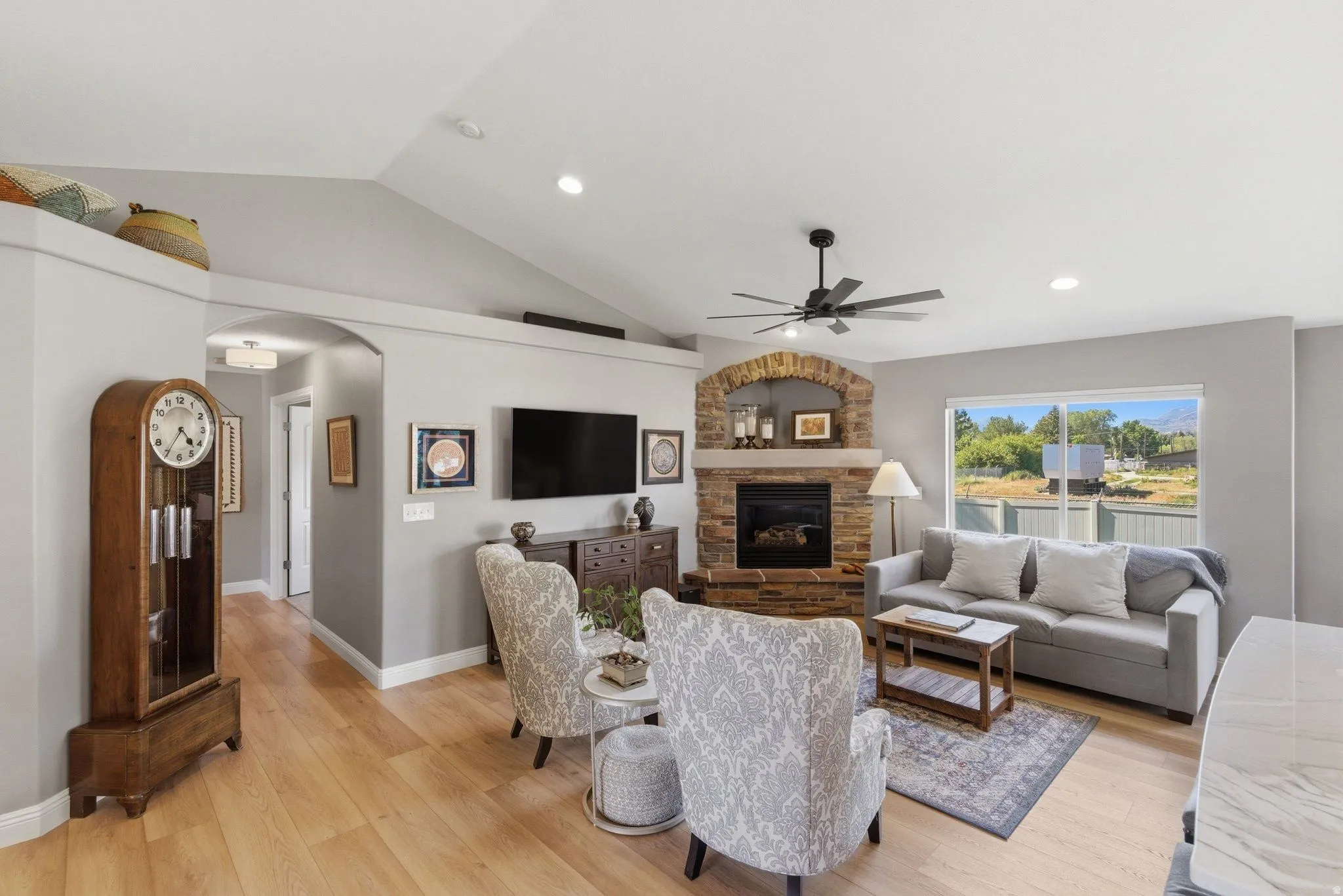 Spacious living area featuring a stone fireplace with a brick arch, light wood-finish flooring, recessed lighting, and a ceiling fan