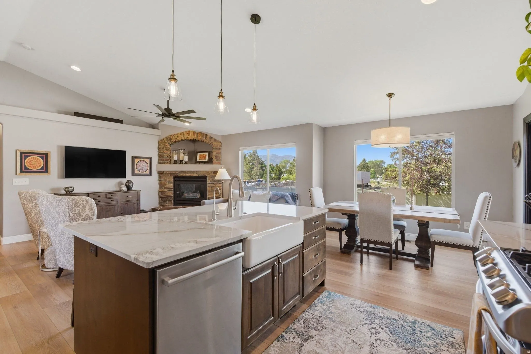 Open-concept interior featuring wood-finish flooring, a kitchen island with a farmhouse sink and light-toned countertop, a stone-accented fireplace, and vaulted ceilings with a ceiling fan