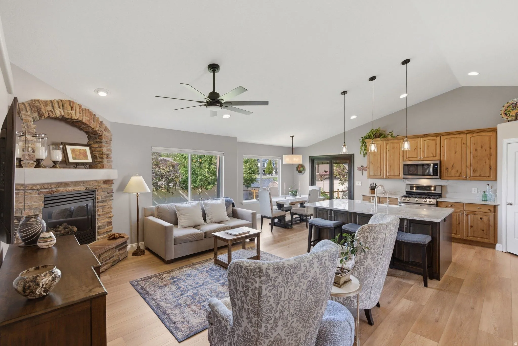 Open-concept living area featuring wood-finish flooring, a stone-clad fireplace with an arched hearth, vaulted ceilings with recessed lighting, and a kitchen island with pendant lighting
