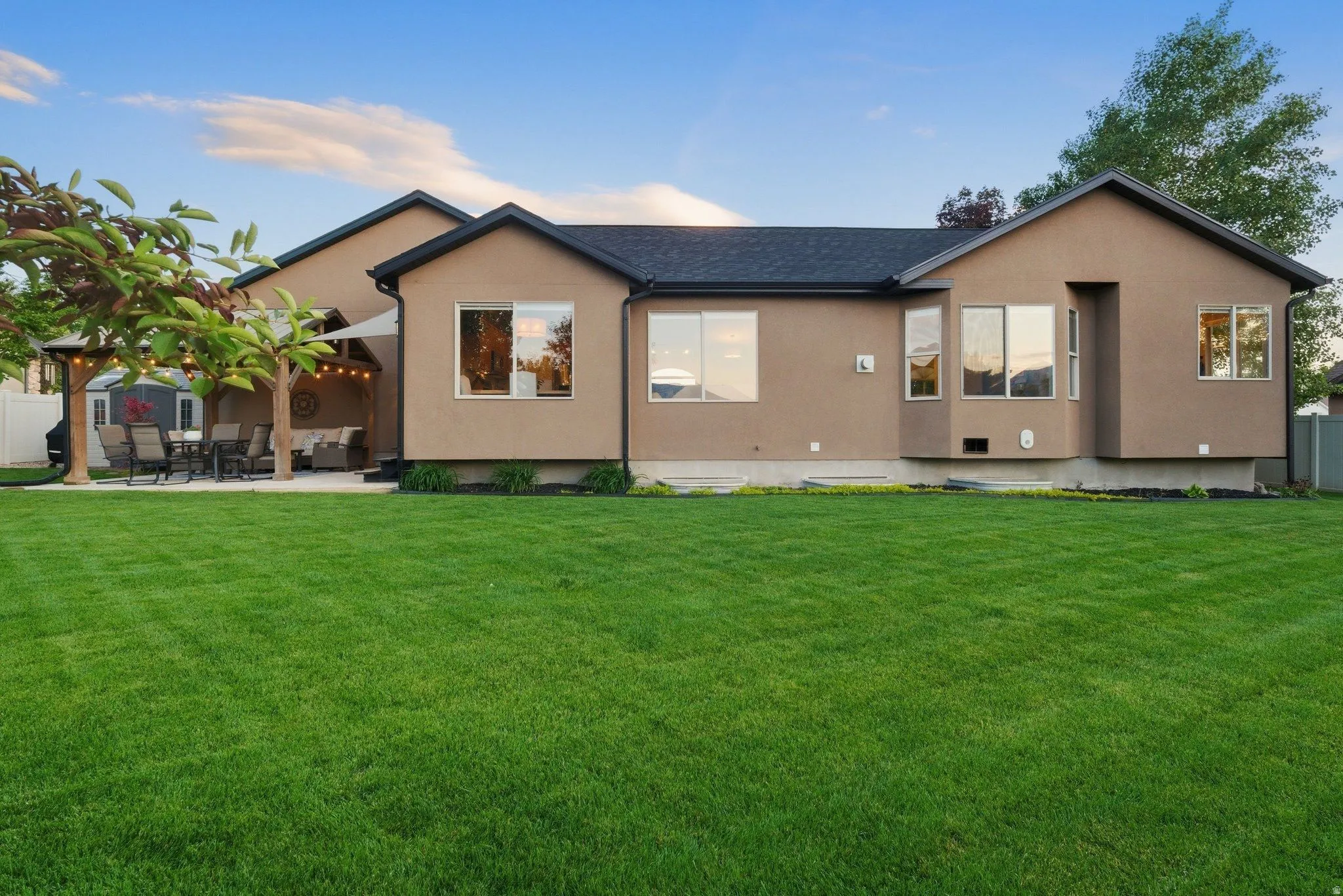 Expansive green lawn with a stucco exterior home featuring multiple windows, a rear patio, and a covered outdoor entertaining area