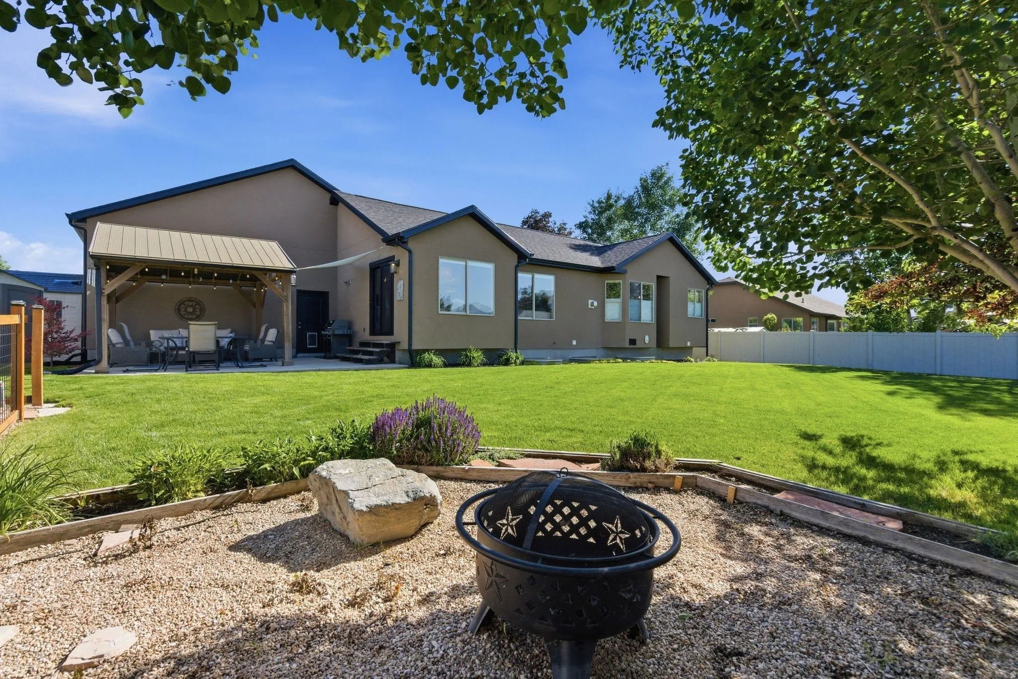 Expansive backyard featuring a manicured lawn, a gravel fire pit area with a decorative fire pit, and a covered patio with string lighting