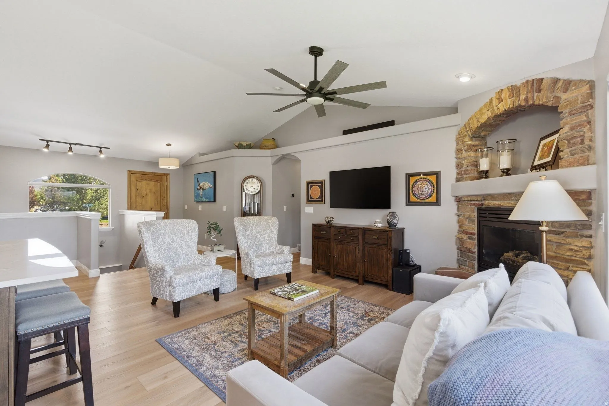 Spacious living area featuring a vaulted ceiling with a ceiling fan, light wood-finish flooring, and a stone-accented fireplace with an arched hearth