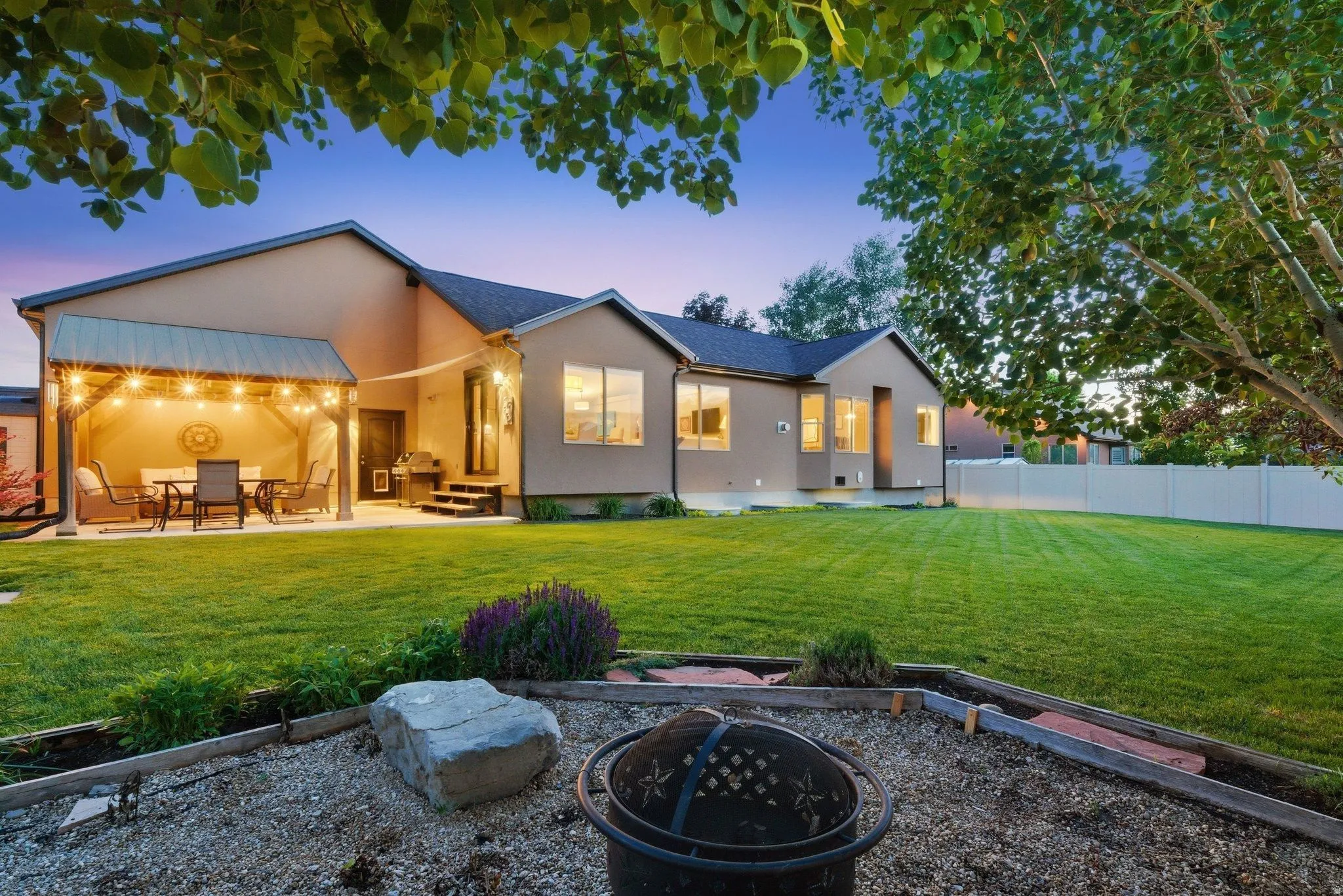 Spacious backyard featuring a covered patio with string lighting, expansive lawn, gravel fire pit area, and a privacy fence