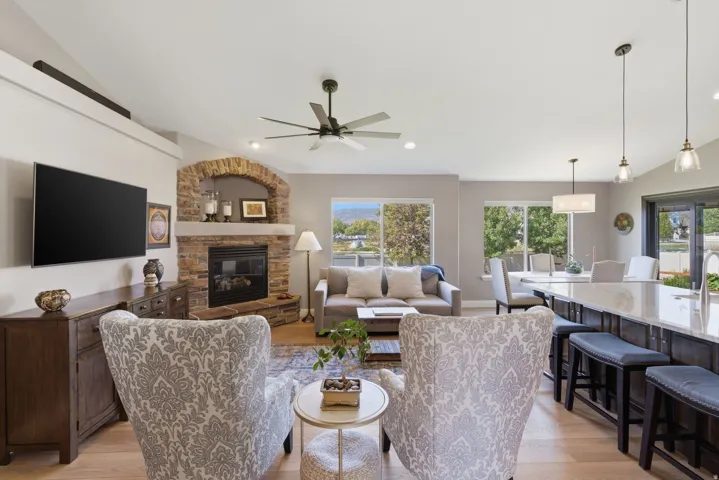 Vaulted ceiling living area featuring a stone-clad fireplace with an arched opening, wood-finish flooring, a ceiling fan, and large windows