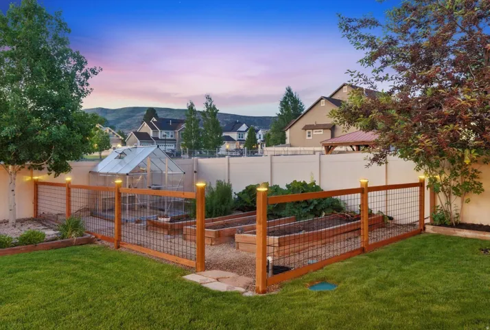 Backyard featuring a functional greenhouse, raised garden beds, wood and wire fence, manicured lawn, and mature trees