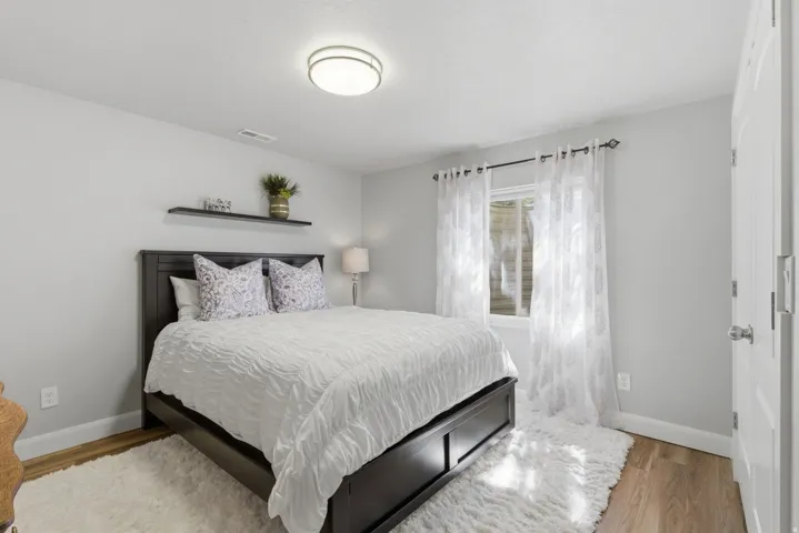 Bedroom featuring wood-finish flooring, light gray walls, a window with sheer white curtains, and a flush-mount ceiling light