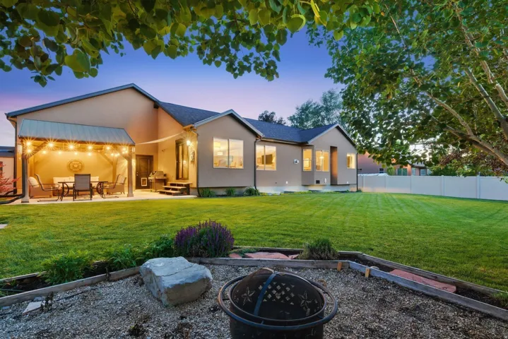 Spacious backyard featuring a covered patio with string lighting, expansive lawn, gravel fire pit area, and a privacy fence