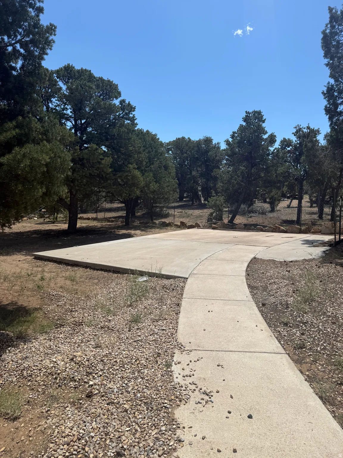 Picnic area concrete pathway leading to a spacious concrete pad, surrounded by extensive natural landscaping with mature trees and a gravel border
