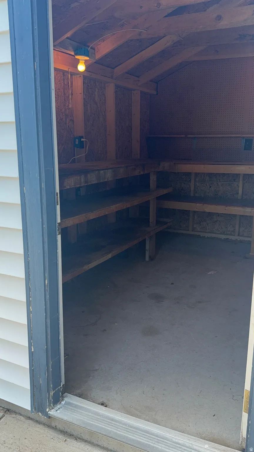 Utility shed interior featuring built-in shelving, concrete flooring, exposed rafters, and an overhead light fixture